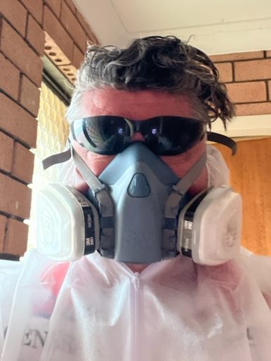**Alt text:** A person wearing a full protective suit, including a respirator mask, sunglasses, and a white hazmat-style coverall. They are standing indoors with a brick wall and a door in the background. 