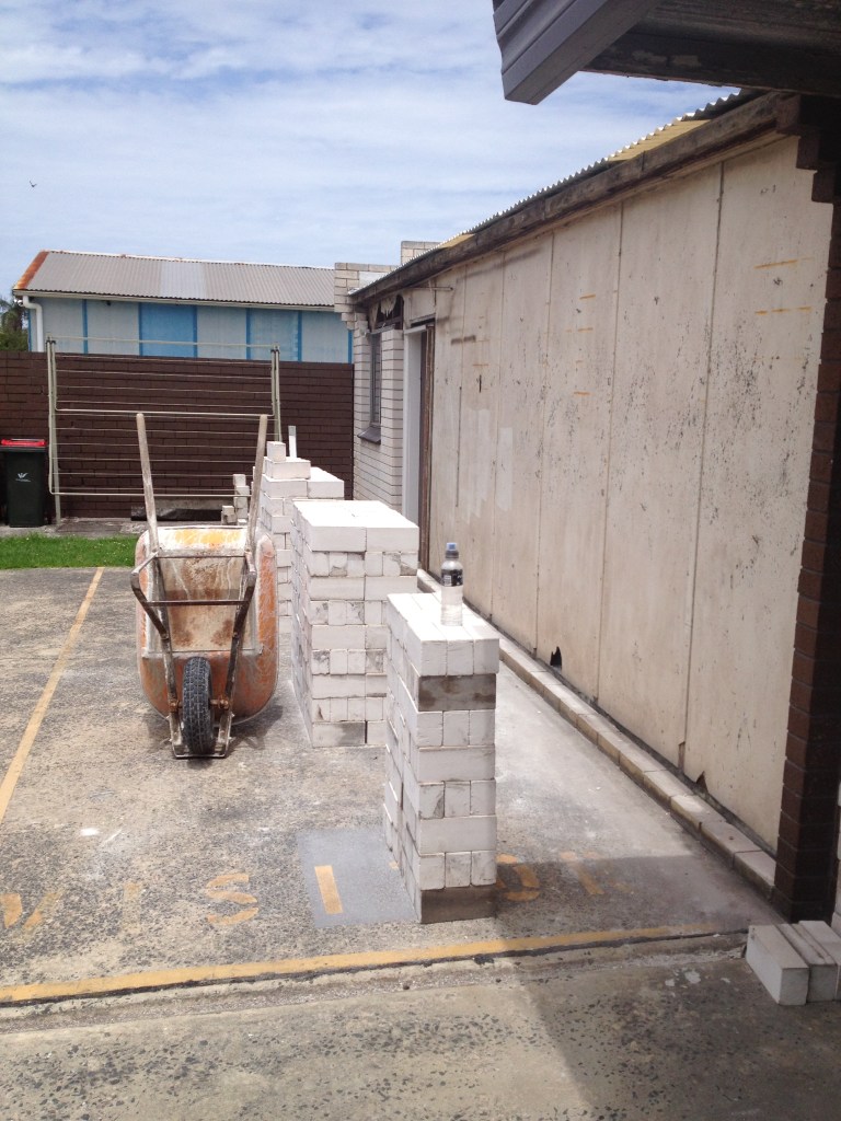 A construction site with a wheelbarrow in front of a partially built brick wall near a residential shed.