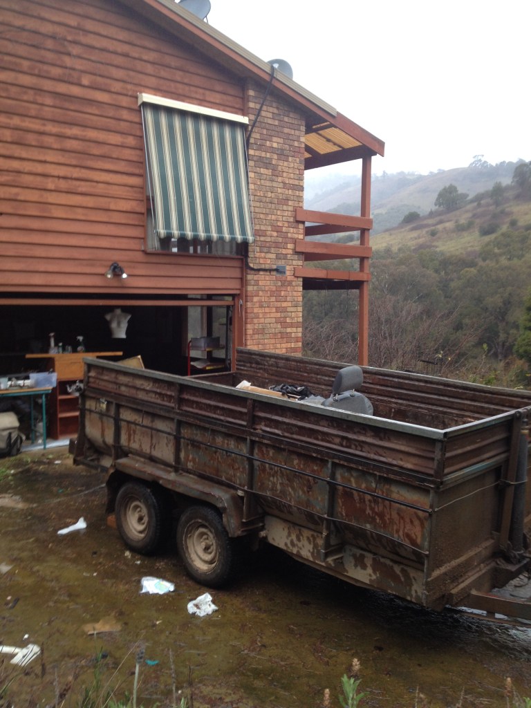  A rusty trailer parked outside a wooden house, with trash scattered around the ground.
