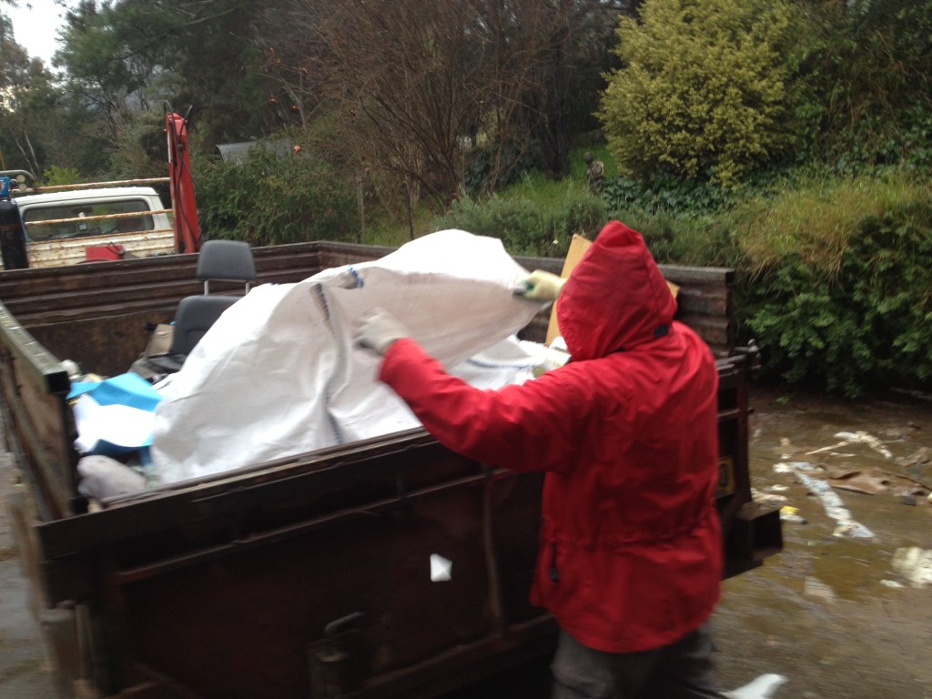A person wearing a red hooded jacket lifting a large white tarp in a trailer filled with waste, with trees in the background.

