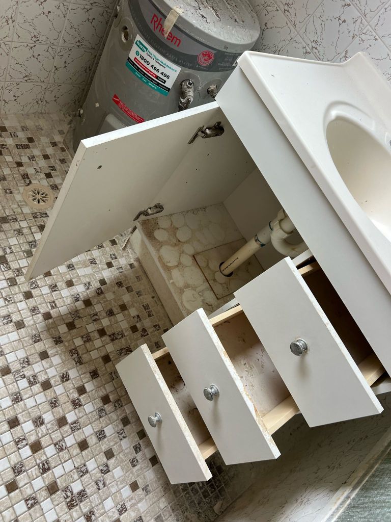 A damaged bathroom vanity with open and broken drawers, exposing plumbing. A small water heater is mounted on the wall in the background. The floor has a mosaic tile pattern.

